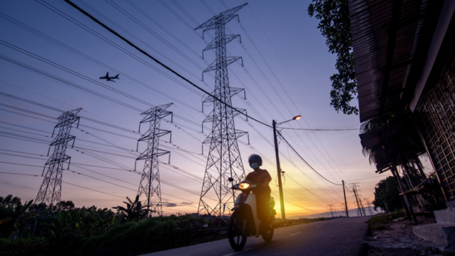 A person riding motorcycle on road in Malaysia at sunset, beside electrical transmission towers with a plane flying overhead. (photo)