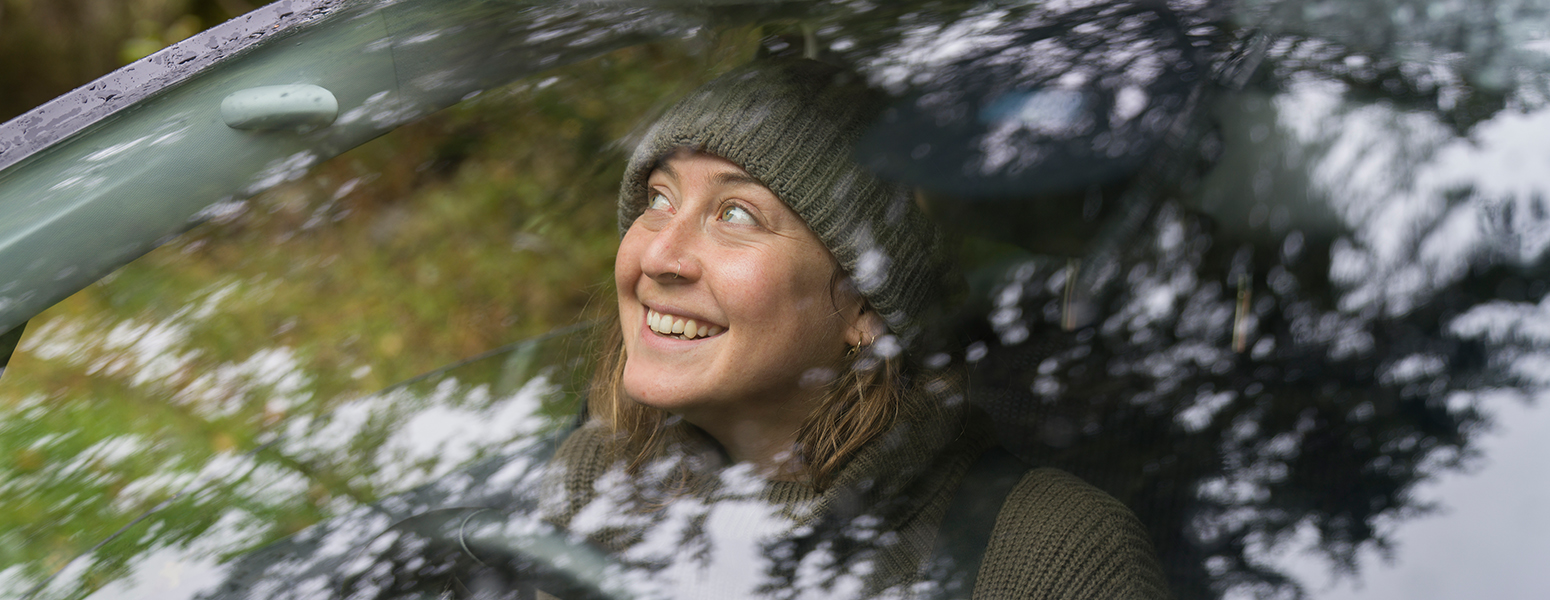 Smiling woman wearing a cap looking out of the front window of a car surrounded by trees (photo)