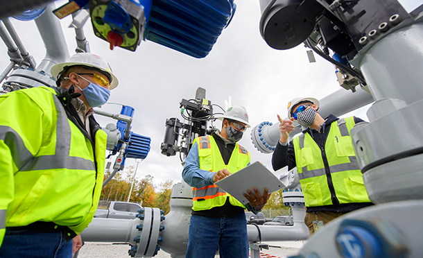 Three men wearing safety equipment and masks discussing safety topics near a pipeline (photo)