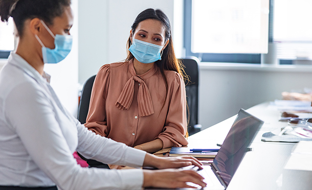 Two woman wearing masks sitting together and looking at a laptop screen (photo)