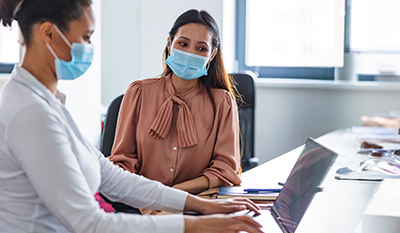 Two woman wearing masks sitting together and looking at a laptop screen (photo)