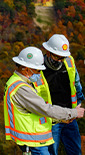Three men wearing safety equipment and masks discussing safety topics near a pipeline which crosses a wood in the background (photo)