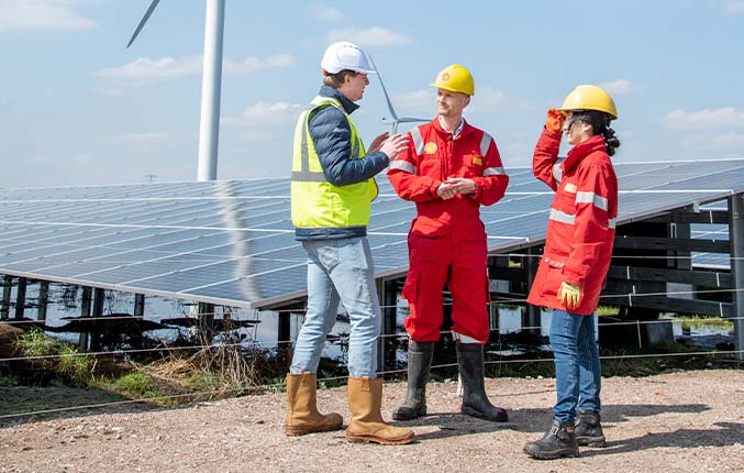 two men and a woman wearing PPE standing together talking next to a solar panel with wind turbine in the background (photo)